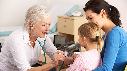 A mother holding a child whilst a GP checks on her using a stethoscope.